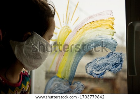 A child in a medical mask looks through the glass to the street. The kid draws a rainbow on a window pane as a symbol of a dream and hope for the future. The kid is sad on the street sitting at home i