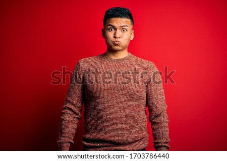 Young handsome latin man wearing casual sweater standing over red background puffing cheeks with funny face. Mouth inflated with air, crazy expression.