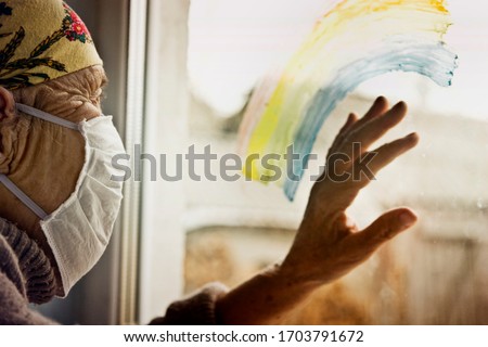 An old grandmother in a protective mask looks out the window at self-isolation. Elderly woman at quarantine at home. Grandmother looks through the rainbow on the window pane into the street with a clo