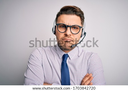 Young call center operator business man with blue eyes wearing glasses and headset skeptic and nervous, disapproving expression on face with crossed arms. Negative person.