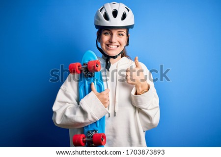 Young beautiful redhead skateboarder woman wearing safety helmet holding skate happy with big smile doing ok sign, thumb up with fingers, excellent sign