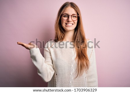 Young beautiful redhead woman wearing casual sweater and glasses over pink background smiling cheerful presenting and pointing with palm of hand looking at the camera.