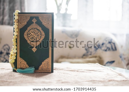 Green covered Muslim Holy book Qu'ran in Arabic "The noble Qur'An" stands on a table against the window, and an onyx rosary hangs on its cover