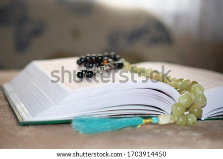 Green covered Muslim Holy book Qu'ran in Arabic "The noble Qur'An" open and lies on a table against the window, and an onyx and opals rosary hangs lie on on its pages