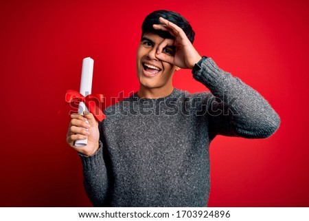 Young student man holding university graduated diploma degree over red background with happy face smiling doing ok sign with hand on eye looking through fingers