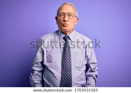 Grey haired senior business man wearing glasses standing over purple isolated background making fish face with lips, crazy and comical gesture. Funny expression.