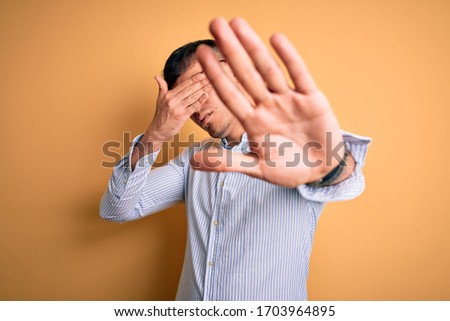 Young handsome hispanic business man wearing nerd glasses over yellow background covering eyes with hands and doing stop gesture with sad and fear expression. Embarrassed and negative concept.