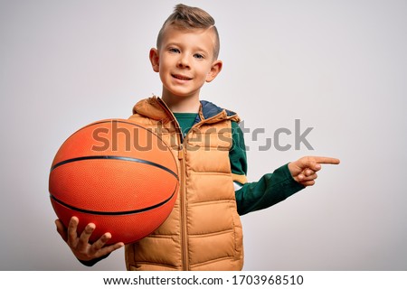 Young little caucasian sports kid playing basketball holding orange ball over isolated background very happy pointing with hand and finger to the side