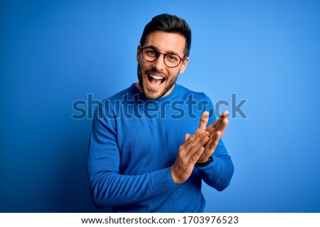 Young handsome man with beard wearing casual sweater and glasses over blue background clapping and applauding happy and joyful, smiling proud hands together Royalty-Free Stock Photo #1703976523