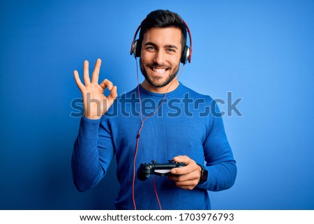 Young handsome gamer man with beard playing video game using joystick and headphones doing ok sign with fingers, excellent symbol