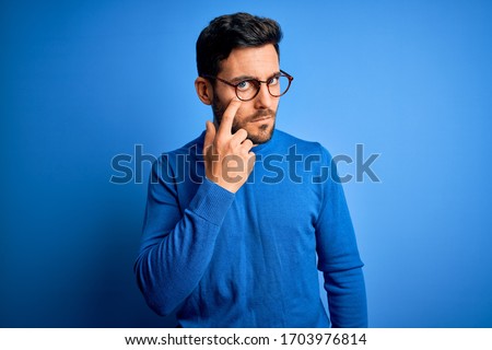 Young handsome man with beard wearing casual sweater and glasses over blue background Pointing to the eye watching you gesture, suspicious expression Royalty-Free Stock Photo #1703976814