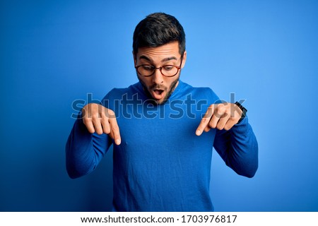 Young handsome man with beard wearing casual sweater and glasses over blue background Pointing down with fingers showing advertisement, surprised face and open mouth
