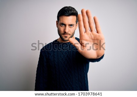 Young handsome man with beard wearing casual sweater standing over white background doing stop sing with palm of the hand. Warning expression with negative and serious gesture on the face.