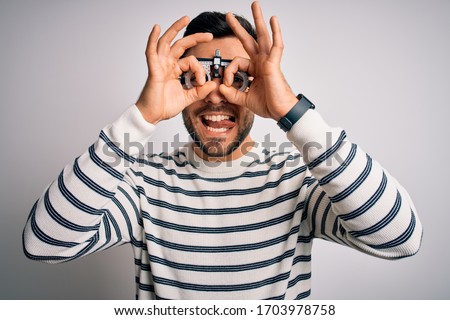 Young handsome man with beard wearing optometry glasses over isolated white background doing ok gesture like binoculars sticking tongue out, eyes looking through fingers. Crazy expression.