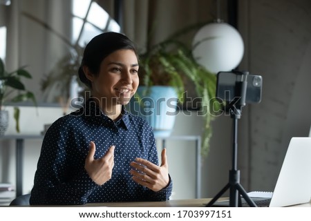 Smiling Indian young woman coach or speaker record live video broadcast on modern smartphone gadget, positive ethnic millennial girl shoot webinar or online training course on cellphone device Royalty-Free Stock Photo #1703997151