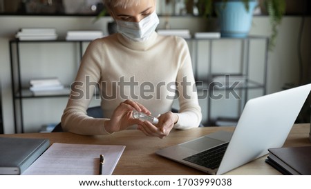 Female employee in protective face mask disinfect hands with sanitizer in office, protect from coronavirus pandemic, woman worker sanitize cleanse use antibacterial gel at workplace, covid-19 concept Royalty-Free Stock Photo #1703999038