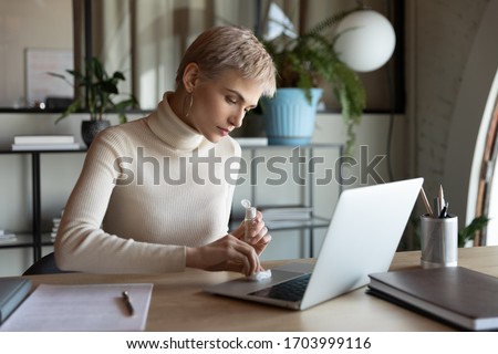 Young businesswoman sit at workplace cleanse laptop keyboard with antibacterial liquid during coronavirus outbreak, female employee disinfect computer with sanitizer in office, covid-19 concept Royalty-Free Stock Photo #1703999116