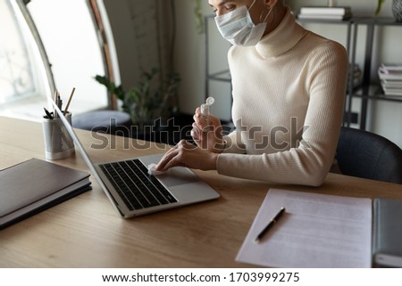 Female employee sit in office wear protective face mask disinfect computer with antibacterial liquid gel protect from coronavirus, woman sanitize laptop workplace with sanitizer from covid-19 pandemic Royalty-Free Stock Photo #1703999275