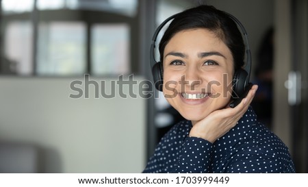 Portrait of smiling Indian young woman in headphones look at camera consult client online, happy ethnic millennial female employee in wireless headset working from home, have web consultation