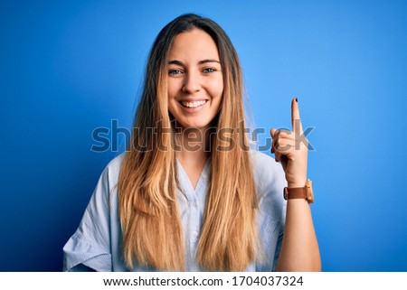 Young beautiful blonde woman with blue eyes wearing striped shirt over blue background pointing finger up with successful idea. Exited and happy. Number one.