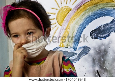 A child in a protective mask is sitting on a quarantine window. A masked kid misses the street due to an outbreak of coronavirus and waits for quarantine to end. Little girl on a background of a rainb