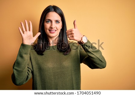 Young brunette woman with blue eyes wearing green casual sweater over yellow background showing and pointing up with fingers number six while smiling confident and happy.
