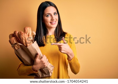 Young woman with blue eyes holding paper bag with bread over isolated yellow background very happy pointing with hand and finger