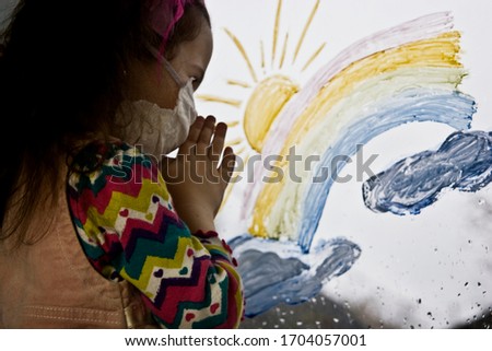 A masked child prays while looking out the window about the end of quarantine and the coronavirus pandemic. The child looks through the rainbow on the window pane into the street with hope.