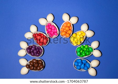 Rabbits made of chocolate eggs and colored jellies form a semicircle on a blue background. Each rabbit coincides with one color of jelly. Photo was taken by flat lay. There is a place for text.