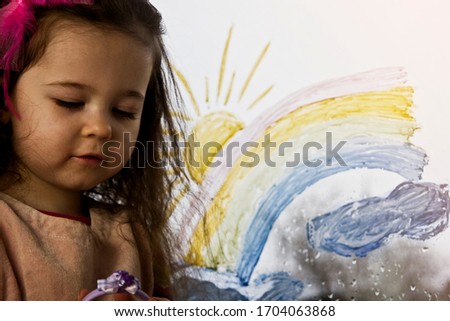 A child sits on a windowsill against the background of a painted rainbow on a window pane in the background. A child painting bright rainbow colors on a glass window during a quarantine attempted for 