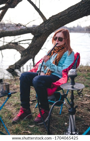 Girl smokes a hookah on the nature. Camping outdoor recreation. Lake. Portable, folding equipment for hiking trips. Lightweight, compact Chair and table made of aluminum tubes. Vertical photo