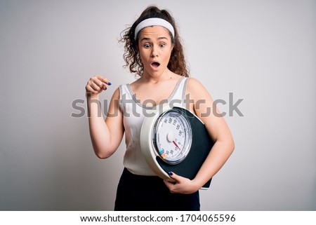 Young beautiful woman with curly hair holding weighing machine over white background Pointing down with fingers showing advertisement, surprised face and open mouth