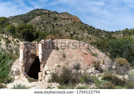 An ancient ice house or snow well with domed roof and arched doorway sits among wild shrubs beneath the rugged mountains near El Garruchal in Murcia, Spain. Blue sky with wispy clouds.