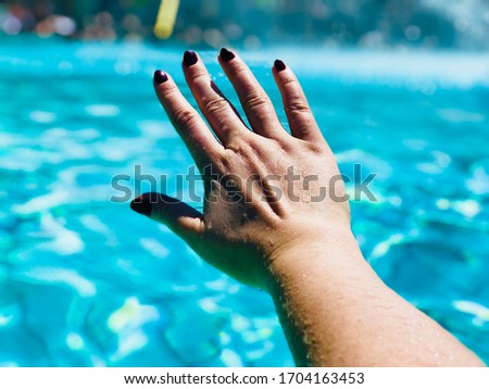 Woman’s hand with dark nail polish in front blue water from a summer pool.