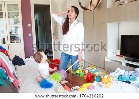 Stressed and tired mother suffering from headache while doing housework with her little baby in messy room. Asian mommy overwork with child. Housewife working Royalty-Free Stock Photo #1704216202