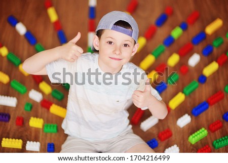 a child a small boy a preschooler in a cap worn backwards sitting on the floor shows a thumbs up-a class sign against the background of colorful blocks of children's construction equipment. childhood,