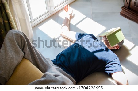 Young man resting on the floor whit a book on his face while enjoying the sun coming through the window. Concept of stay at home, freedom, boredom... Royalty-Free Stock Photo #1704228655