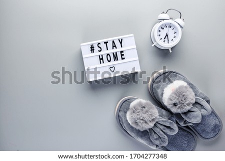 Light box with text STAY HOME, alarm clock and slippers of gray fur with bunnies on gray background. Quarantine and isolation concept. concept. Covid-19, 2019-nCoV. Top view.