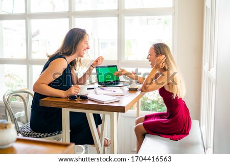 Two young women laughing while working together in a cafe with tea, notebooks and laptop with green screen