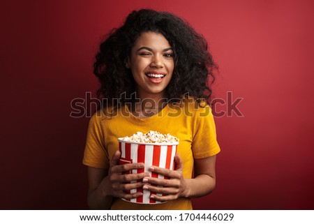 Image of beautiful brunette african american woman with curly hair smiling and holding popcorn bucket isolated over red background