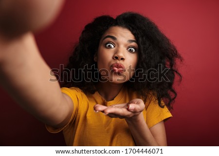 Image of beautiful brunette african american woman sending air kiss while taking selfie photo isolated over red background