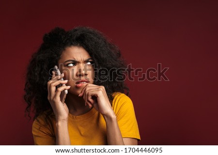Image of displeased brunette african american woman with curly hair expressing outrage while talking on cellphone isolated over red background