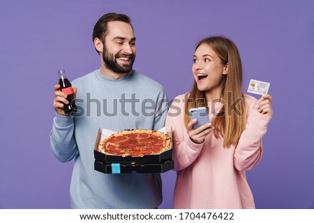 Photo of a cheery emotional loving couple friends isolated over purple wall background drinking soda eat pizza holding credit card using mobile phone.