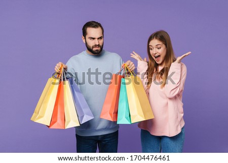 Image of a positive girl near displeased boyfriend isolated over purple wall background holding shopping bags.