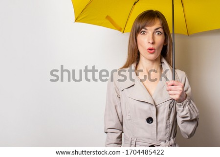 Surprised shock young woman holds a yellow umbrella and looks away against on a light background. Concept rain, bad weather