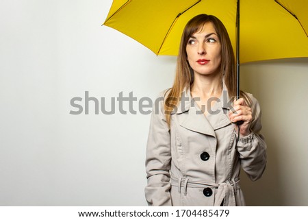 Portrait of a young friendly woman in a classic jacket under a yellow umbrella on an isolated light background. Emotional face. Bad weather, rain, weather forecast