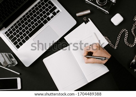 Designer hand writes in his daily planner. Workplace with laptop, vintage film camera, pafume, sunglasses and smartphone on the black desk. Flat lay, top view