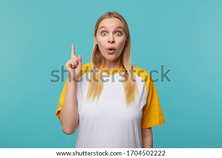 Dazed young attractive fair-haired female with natural makeup rounding surprisedly her blue eyes while raising hand with idea gesture, isolated over blue background