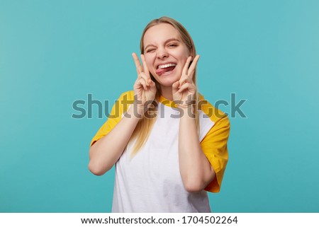 Funny shot of young attractive long haired blonde lady fooling and showing tongue while looking at camera, showing victory sign while standing over blue background