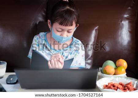 Asian child girl studying homework and wearing face mask during her online lesson at home for protect 2019-nCoV or Covid 19 virus , online education concept.